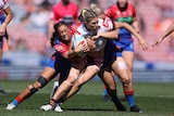 An NRLW player in white carries the ball, with two opponents in blue and red tackling her around the body