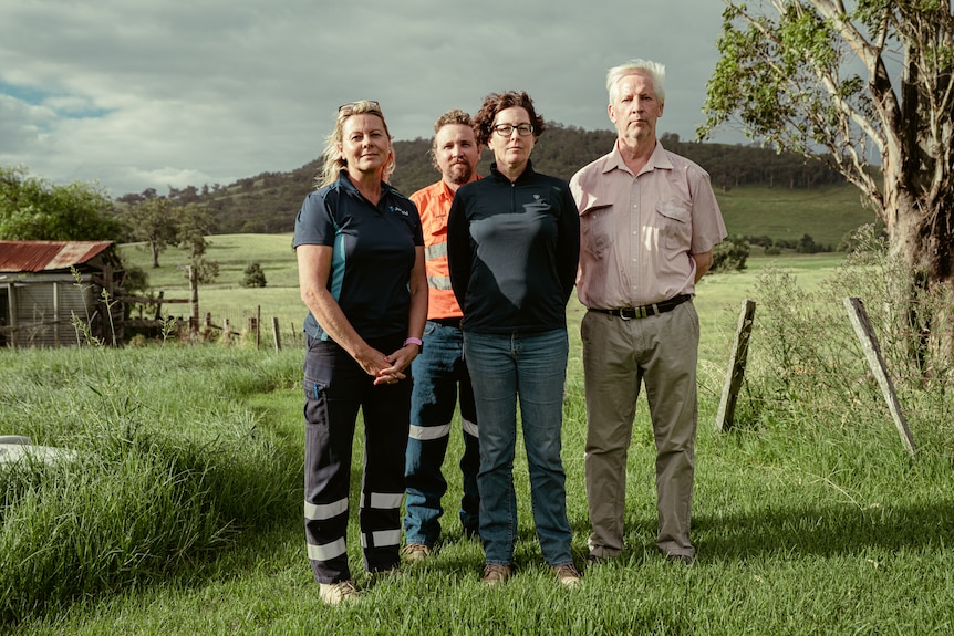 Four people with serious expressions look at the camera standing on a farm