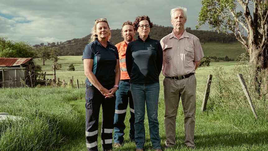 Four people with serious expressions stand on a farm.