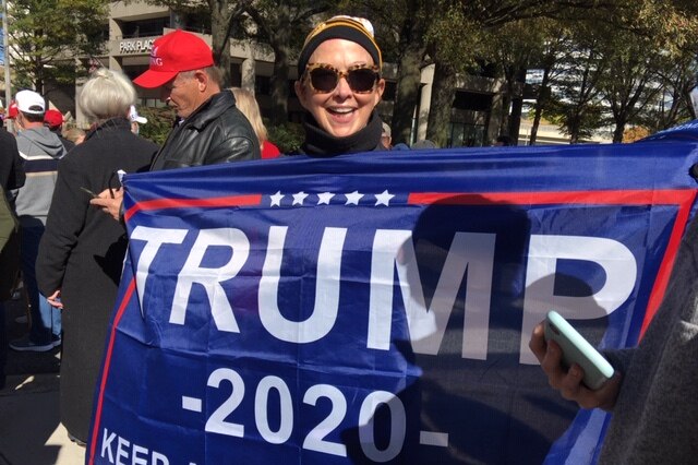 A woman wearing sunglasses and a beanie holds a Trump flag as she smiles in a crowd.