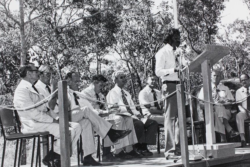 A black and white, historical photo of a Gumatj leader Galarrwuy Yunupingu speaking at a lectern, in front of a row of men.