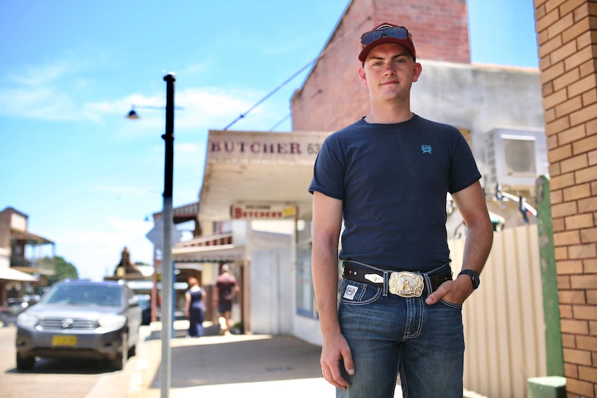 A young man wearing a hat, t-shirt and jeans stands on the street in a small country town.