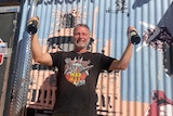 Man wearing Mad Max t-shirt and holding up bottles of champagne standing in front of corrigated iron.