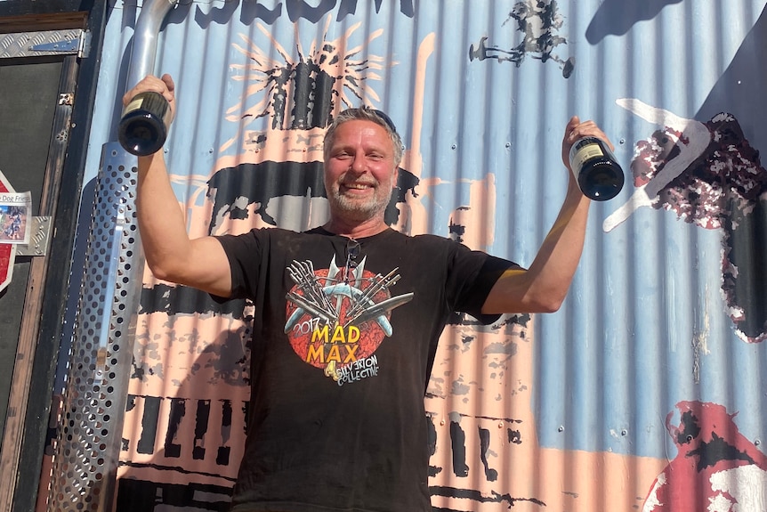 Man wearing Mad Max t-shirt and holding up bottles of champagne standing in front of corrugated iron.