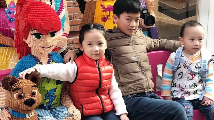 A girl and two boys sit on a bench surrounded by Leggo life size dolls