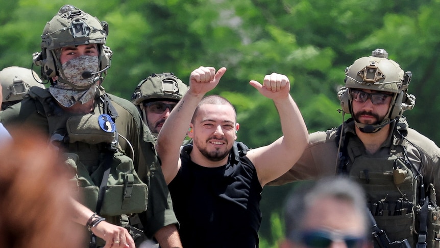 A man with a shaved head is smiling and with his thumbs up, being escorted by two military men.