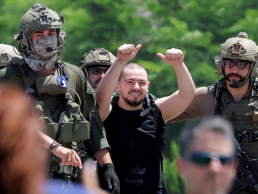 A man with a shaved head is smiling and with his thumbs up, being escorted by two military men.