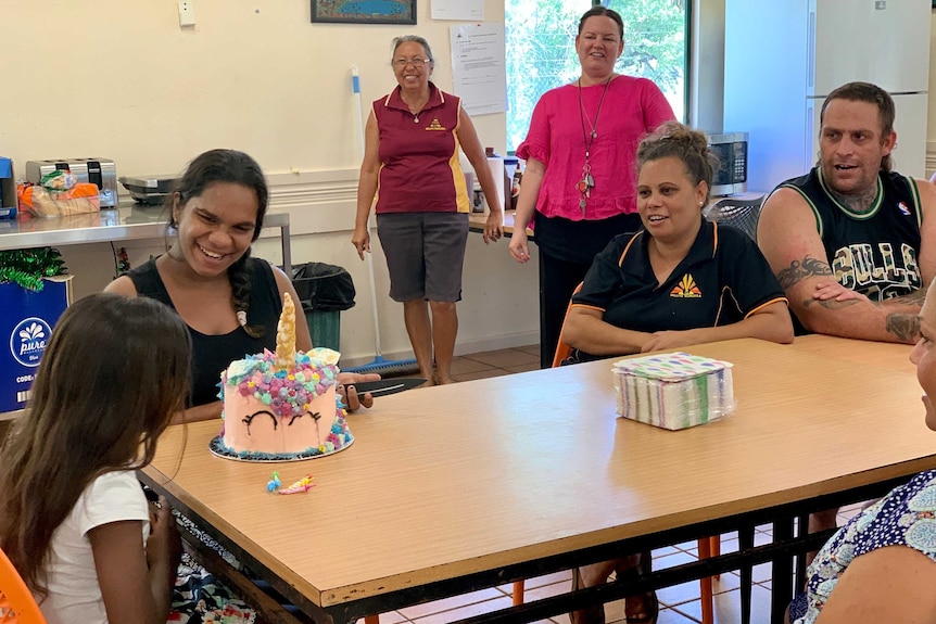 A group of adults and children sit around a table in a sunny room, while a young girl looks at her birthday cake.