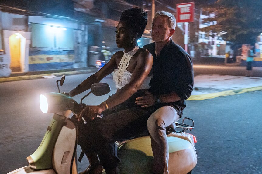 A black woman with dark hair braided into an up-do rides a motorbike with white man with short blonde hair sitting behind her.