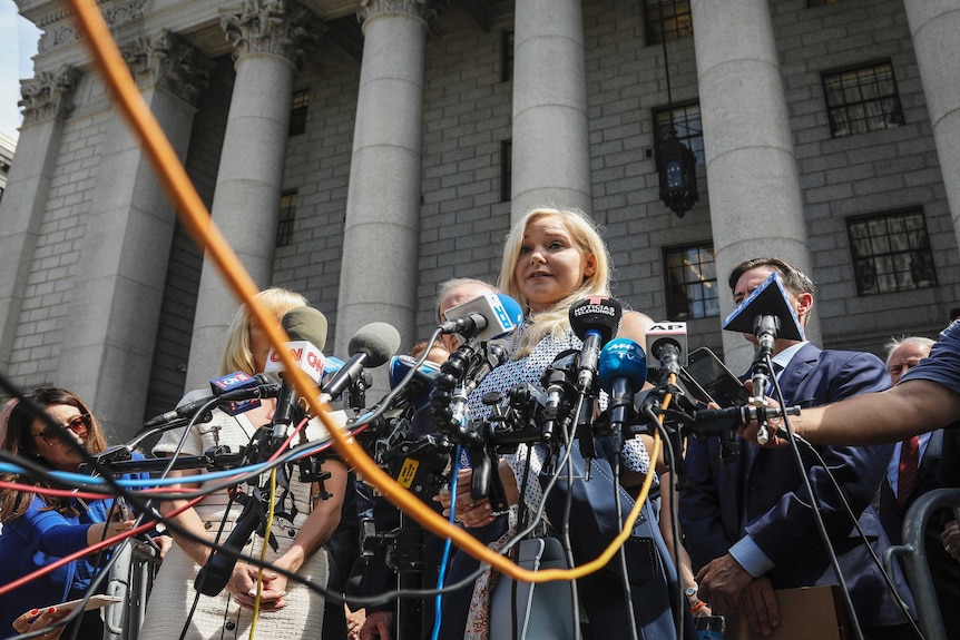 A woman is photographed from a low angle speaking to media behind a large cluster of microphones and wires.