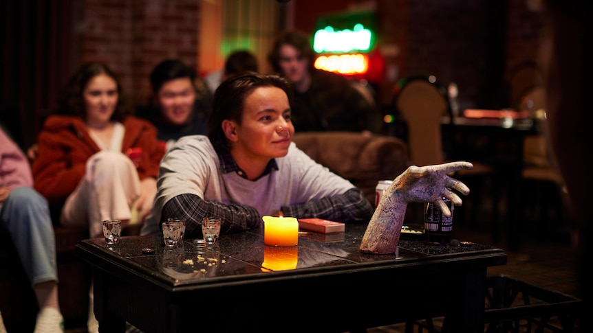 Zoe Terakes sitting at a table with an embalmed hand, people in the background