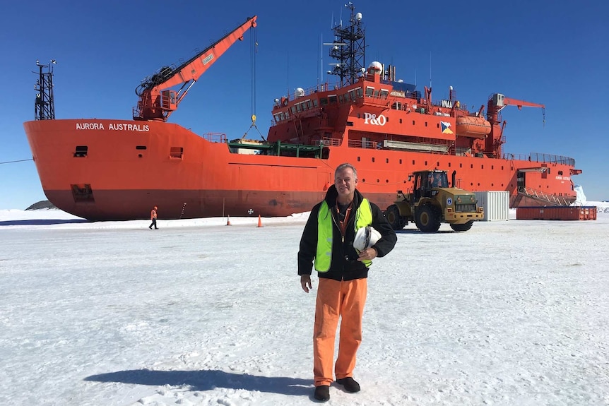 Man stands on ice with a tractor behind him and a large red vessel behind that