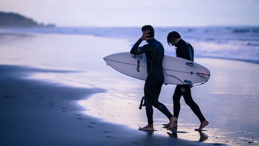 Two men walk along the beach carring their surfboards