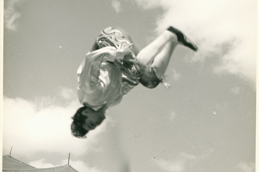 A black and white photo of a man in a costume doing a somersault above a highwire.