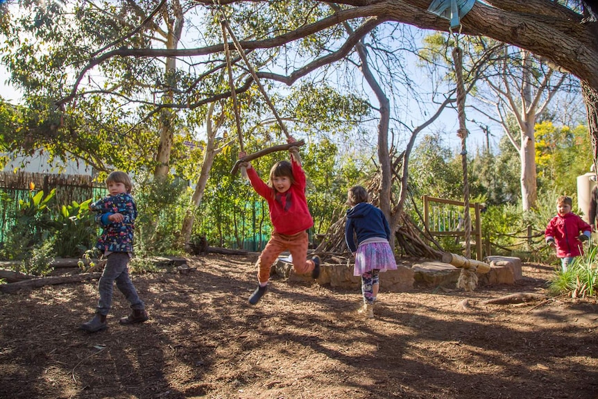 A girl in red hangs off a home-made swing, as two other children play nearby.