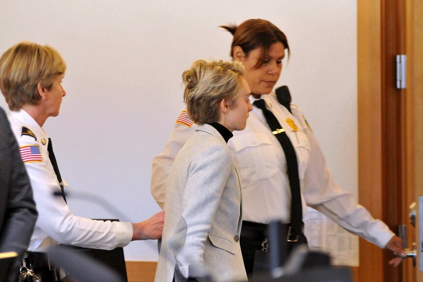 A woman with short blonde hair is escorted by two court officers in uniform.