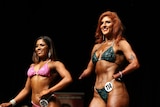 Woman in a green bikini competes in a body building fitness competition on stage. She has no hands. A second woman stands in 