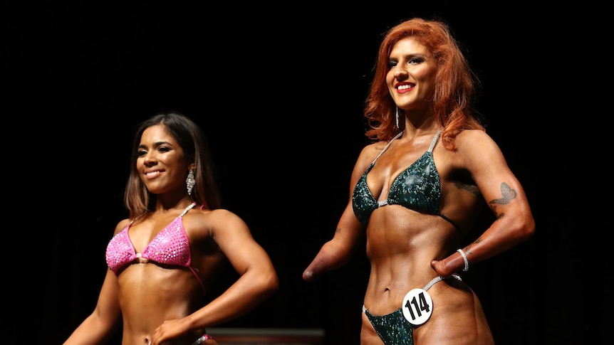 Woman in a green bikini competes in a body building fitness competition on stage. She has no hands. A second woman stands in 