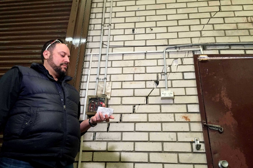 A man stands in front of a brick wall holding a piece of brick.