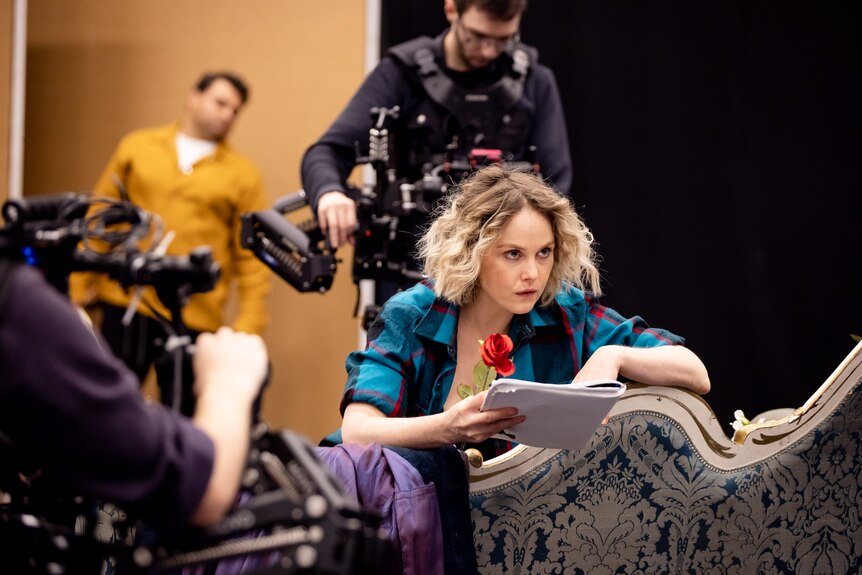 Woman with short curly blonde hair wears blue checked shirt and holds rose and script on settee while two camera operators film