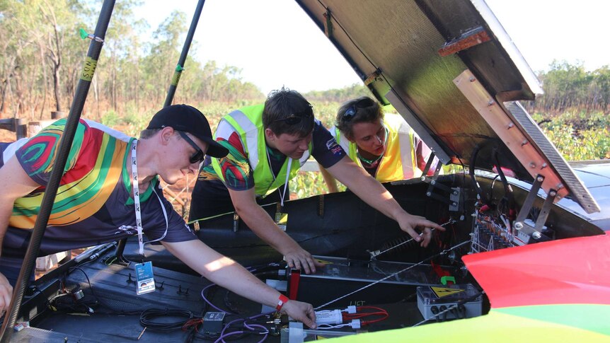 Three lean over and look in the boot of a solar car where there are high voltage batteries with bush in the background.