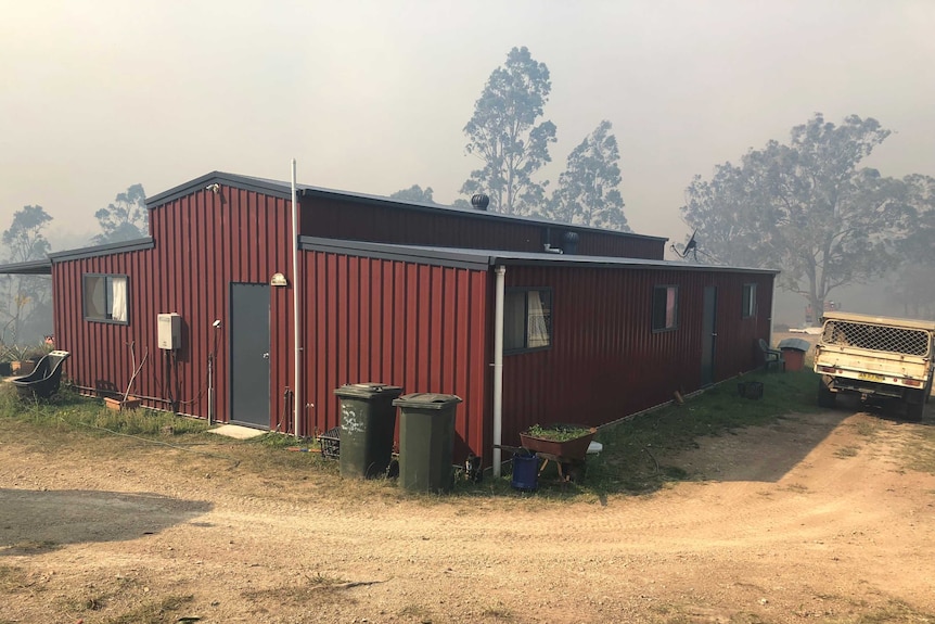 A maroon shed with a ute parked on dirt nearby against a backdrop of tall trees shrouded in smoke.