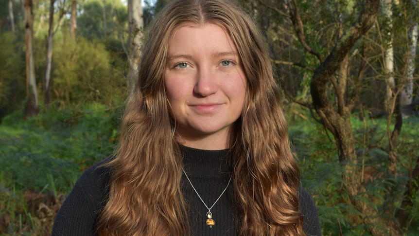 Katta O'Donnell wears a black turtleneck and long hair, standing in a forest in a portrait.