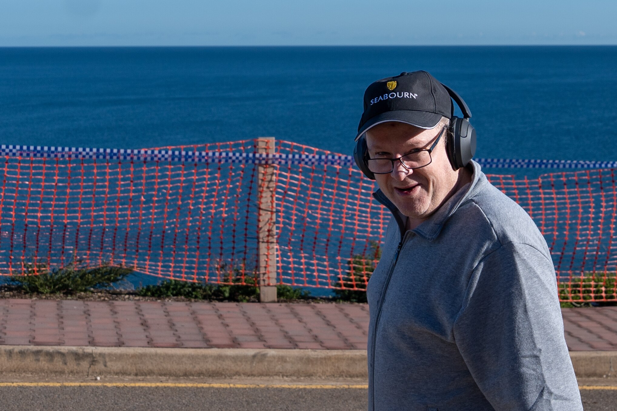 A man wearing glasses, a cap and headphones stand next to police strap and orange netting set up as temporary fence