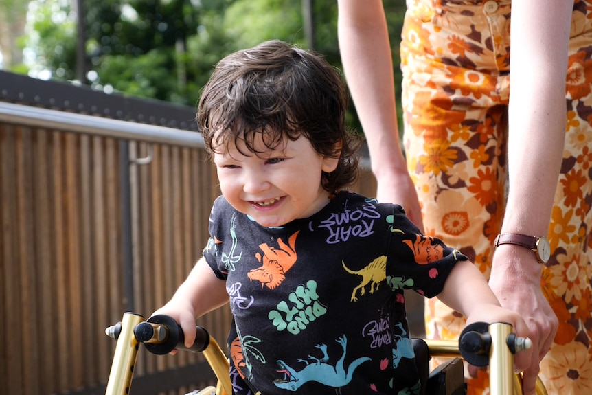 Willow, three-year-old boy smiling and pushing a walker, person behind him.