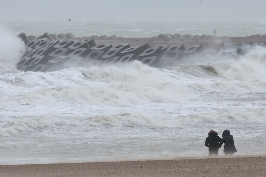 two hooded figures stand at a windy beach 