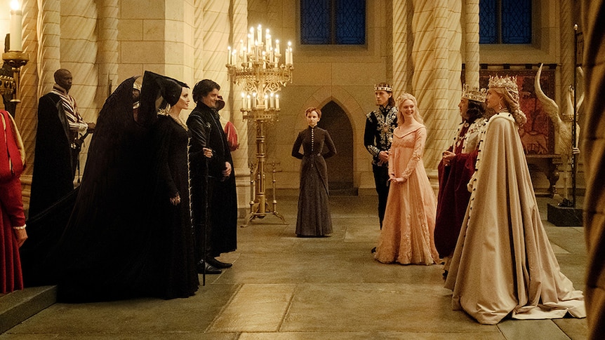 Angelina Jolie and Sam Riley dressed in black stand and face Elle Fanning and Michelle Pfeiffer in large medieval hall.