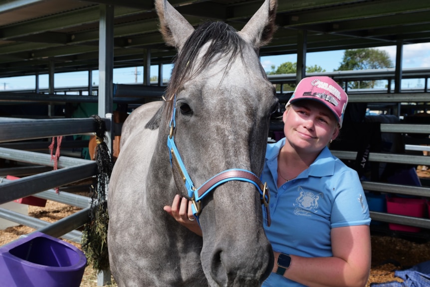 A woman in a blue polo neck wears a pink cap, holds a grey horse in a stable and smiles looking away from the camera.