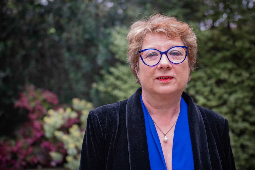 A woman with a dark blue velvet jacket, blue shirt, blue rimmed glasses and short hair, green bushes blurred behind.