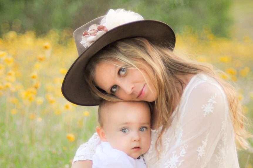 A woman holding a baby sits in a field and looks at the camera.