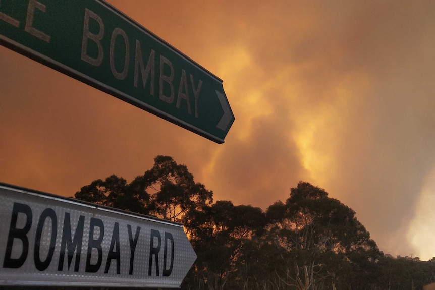 Smoke and a glow from a large fire with a street sign for Little Bombay and Bombay Road in the foreground.