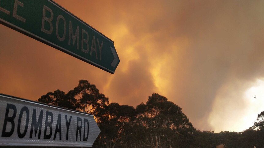Smoke and a glow from a large fire with a street sign for Little Bombay and Bombay Road in the foreground.