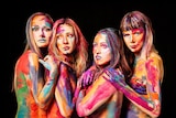 Four female singers in body paint and nothing else huddle together.