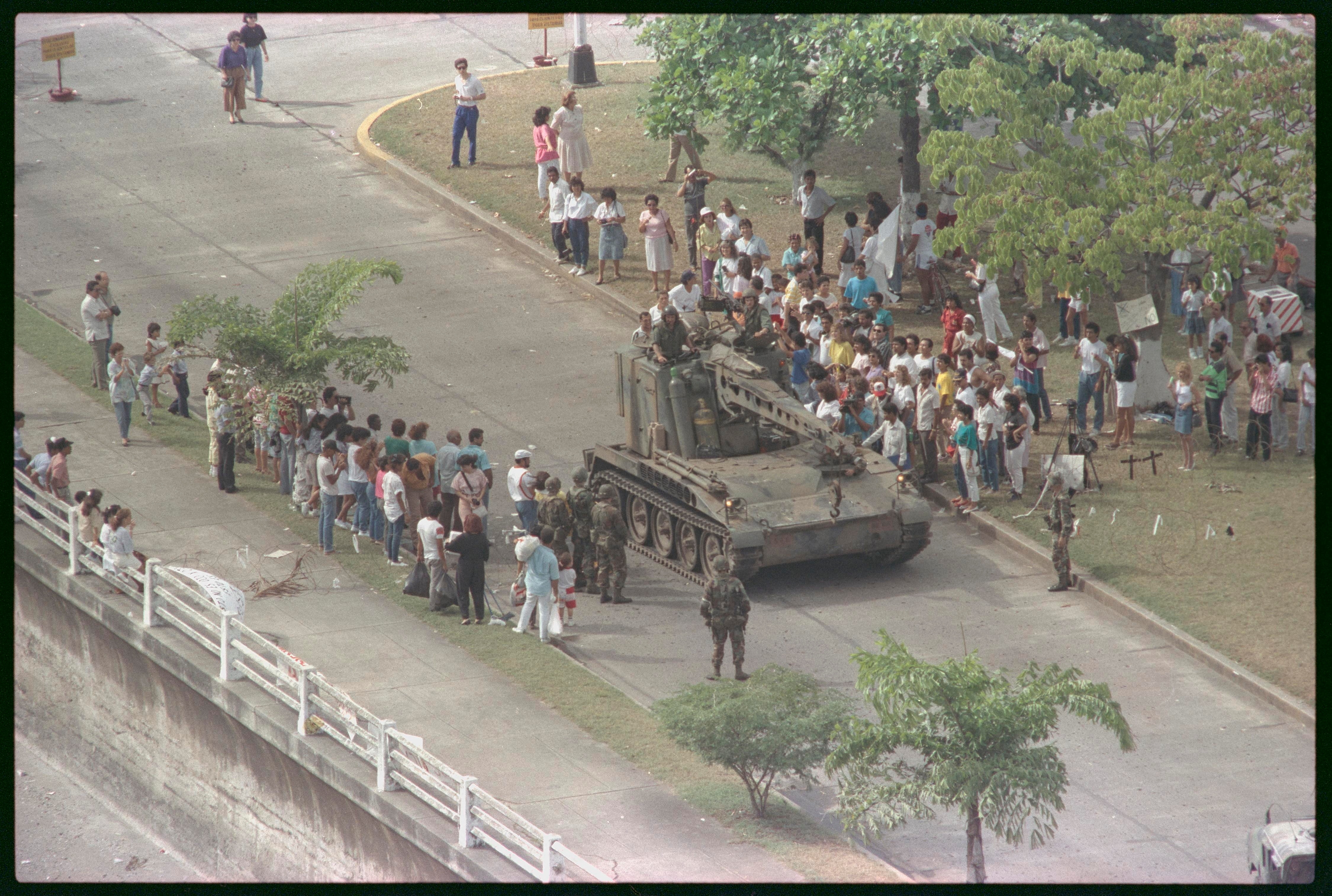 Um tanque rola pela rua enquanto as pessoas se aglomeram dos dois lados. 