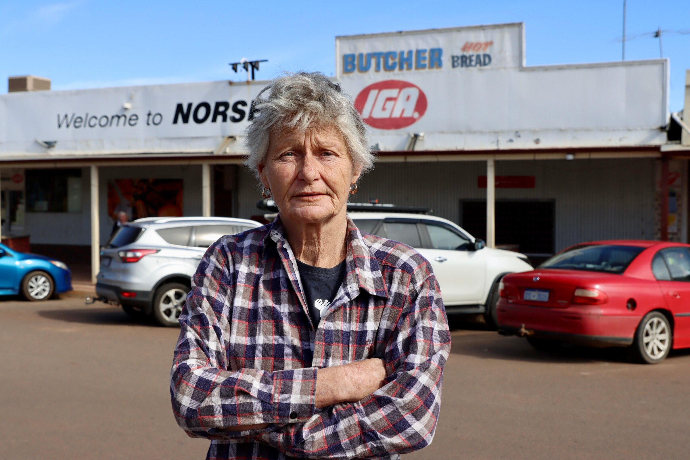 An elderly woman with grey hair standing in main street of country town with arms crossed.  