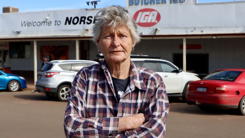 An elderly woman with grey hair standing in main street of country town with arms crossed.  