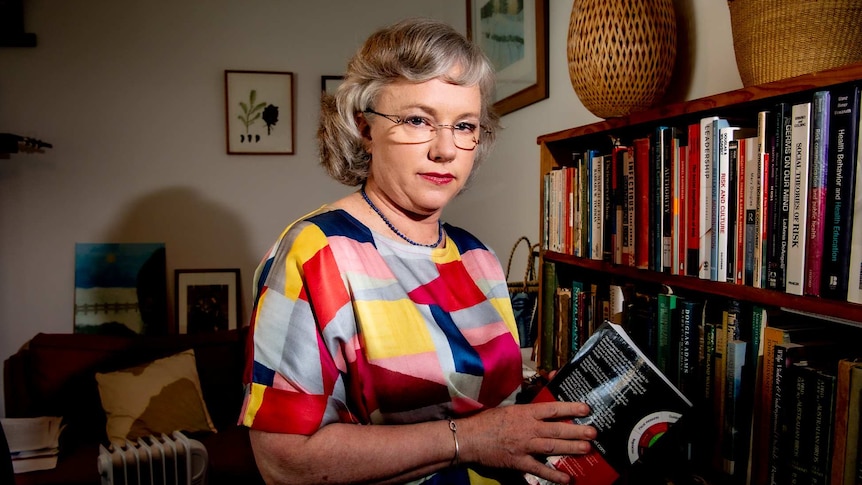 A woman in a colourful top looking down the barrel of the camera near a book shelf