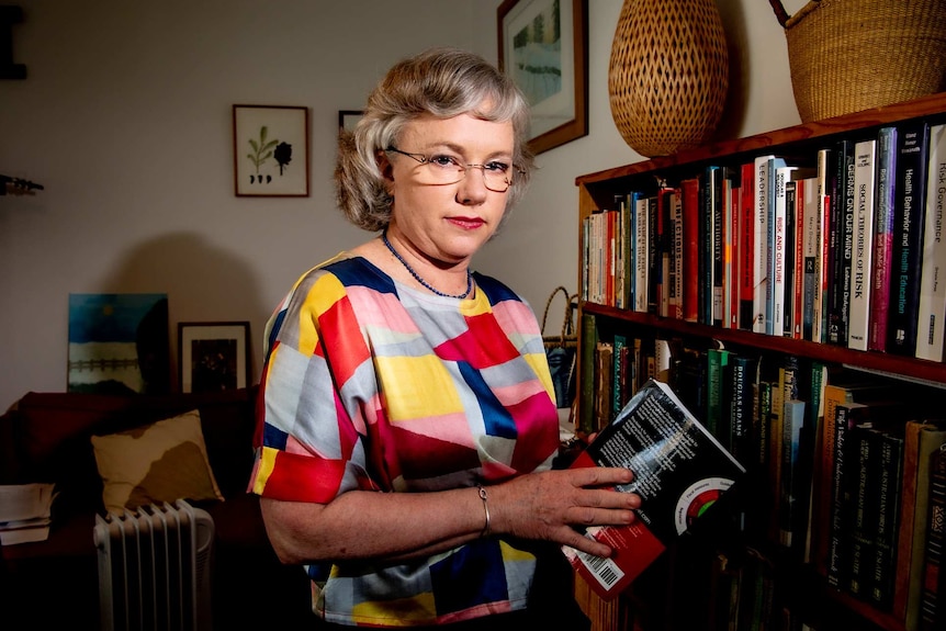 A woman in a colourful top looking down the barrel of the camera near a book shelf