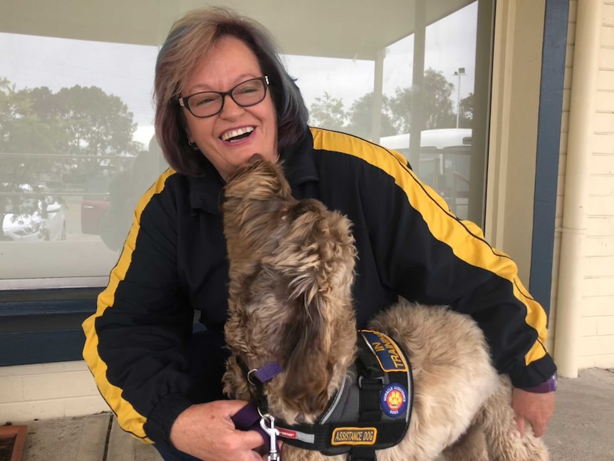 A woman crouched on the ground with an assistance dog licking her face