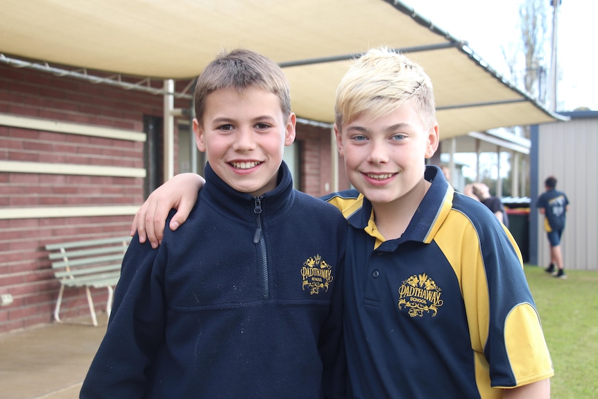 Two boys smile at the camera, wearing school uniforms, with a brick building behind them