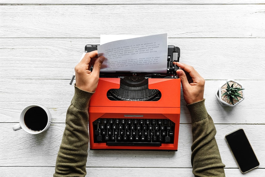 Colour aerial view of hands pulling paper from red typewriter on a white wooden desk.