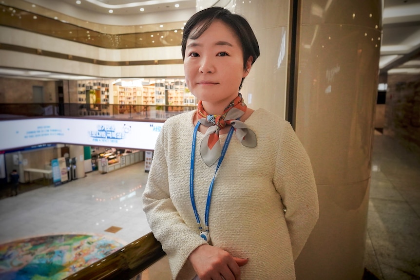 A Korean woman with cropped hair, a cream top and neck scarf leans on a railing in a shopping mall 