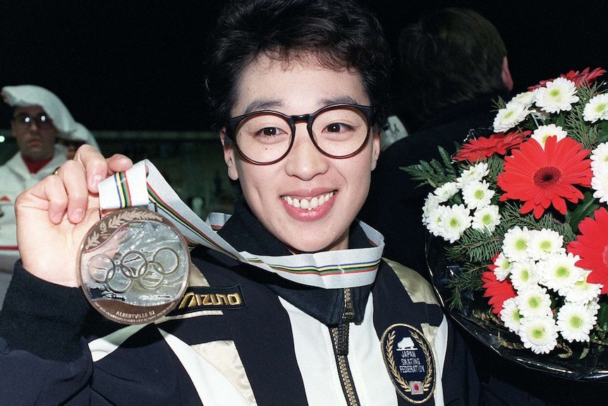 A woman in a track suit smiling while holding a bronze medal and a bunch of flowers
