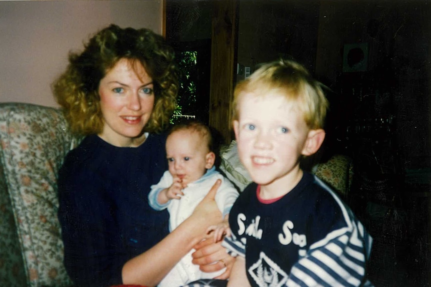 Sam Drummond as a young boy is standing in front of his mum who is cradling his baby brother Jesse in her arms
