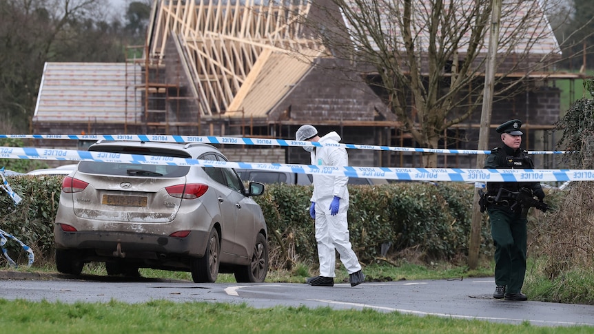 Police tape in front of a car and one person in PPE and another person in a police uniform