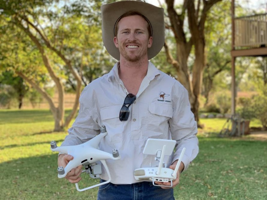 A young man with a broad-brimmed hat smiles as he holds two drones.